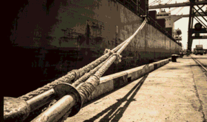 Duracordix - Votre fournisseur de confiance de cordages et filets synthétiques haute performance | Qu'est-ce qu'un cordage d'amarrage ? Guide maritime complet ! Une photographie saisissante en plongée, aux tons sépia, prise depuis un quai portuaire. On y voit un câble d'amarrage synthétique DURACORDIX, robuste et tendu, fixé autour d'une borne d'amarrage. Ce câble épais remonte vers la coque d'un imposant cargo à quai, soulignant sa solidité et sa durabilité.