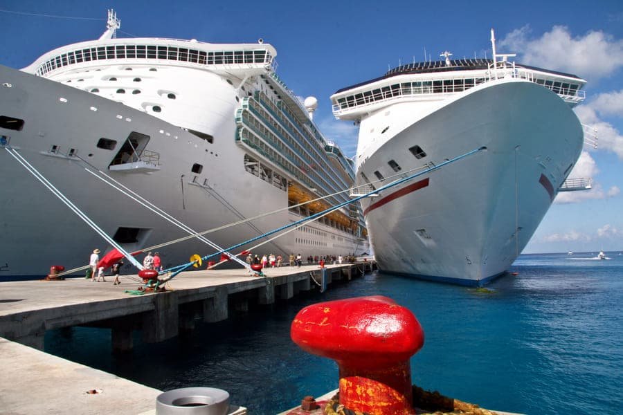 Dois grandes navios de cruzeiro atracados em um porto ensolarado, presos por cabos de amarração a um cais, com o mar azul e um poste de amarração vermelho em primeiro plano.