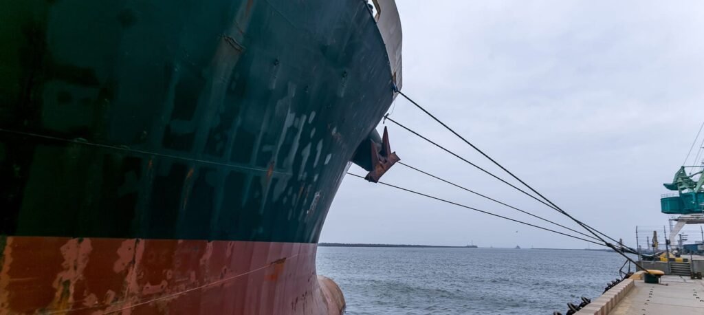 Large cargo ship docked at a port with several mooring lines stretching from the ship’s bow to a bollard on the quay