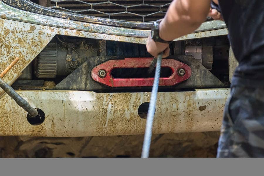 une personne tirant un câble de treuil synthétique UHMWPE bleu haute résistance à travers un guide-câble en aluminium rouge monté sur le pare-chocs boueux d'un véhicule 4x4 lors d'une opération de sauvetage hors route