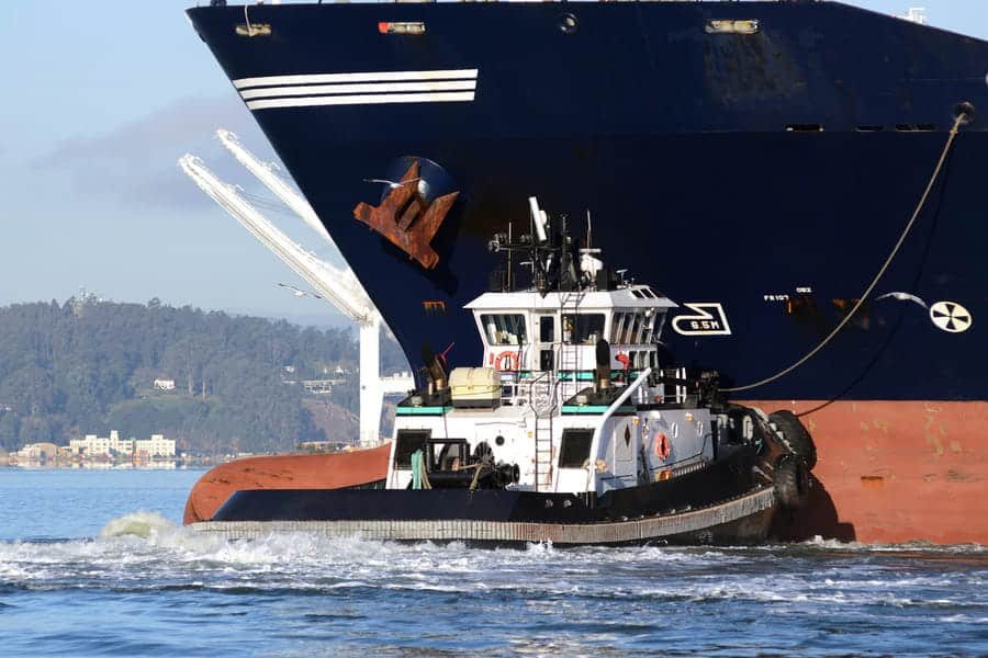 Un remorqueur portuaire assiste un immense cargo en appuyant son étrave contre la coque du navire, déployant des câbles de remorquage marins à haute résistance pour des manœuvres précises dans les eaux calmes du port.